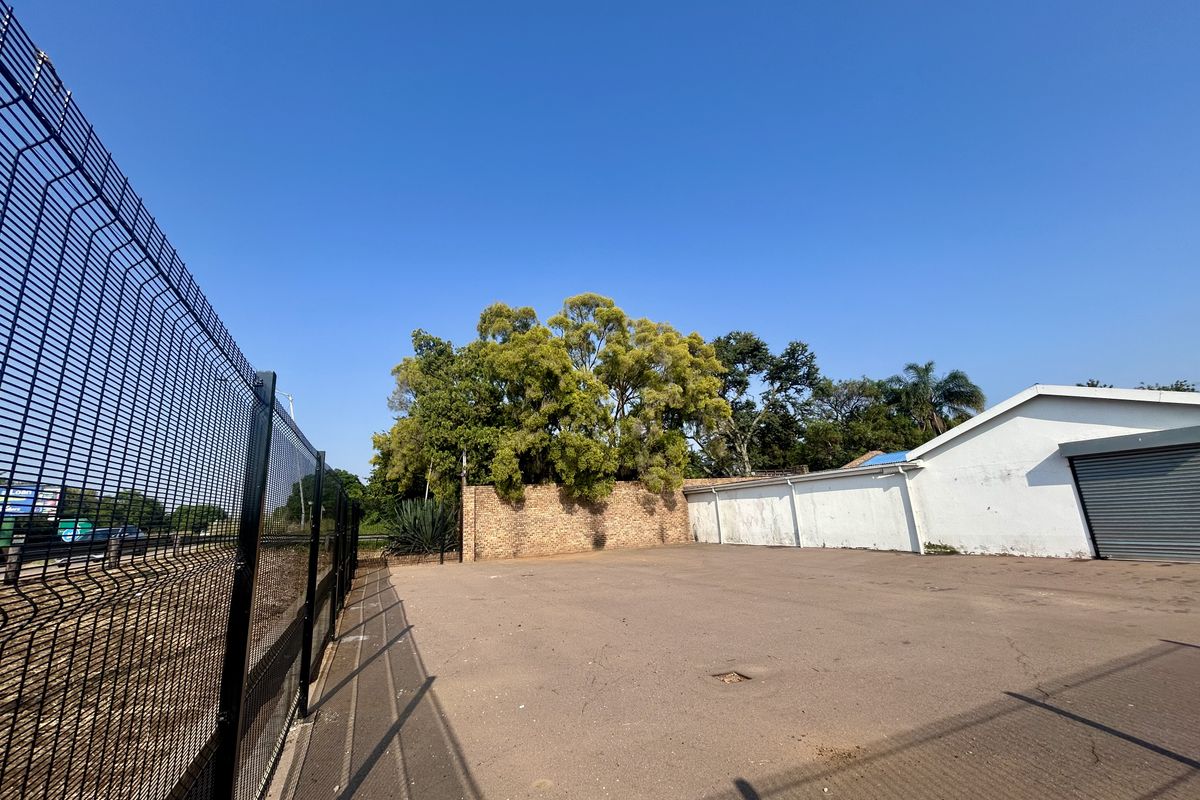 A paved industrial yard area featuring a brick boundary wall, perimeter wire fencing, and a roller shutter door access point.