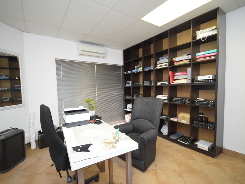 A functional office space featuring a large built-in shelving unit, tiled flooring, suspended ceiling, and a wall-mounted air conditioning unit.