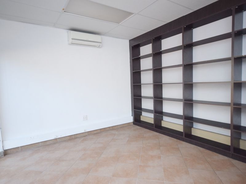 A bright, empty office space featuring tiled flooring, a large built-in dark wood shelving unit, and a wall-mounted air conditioning unit.