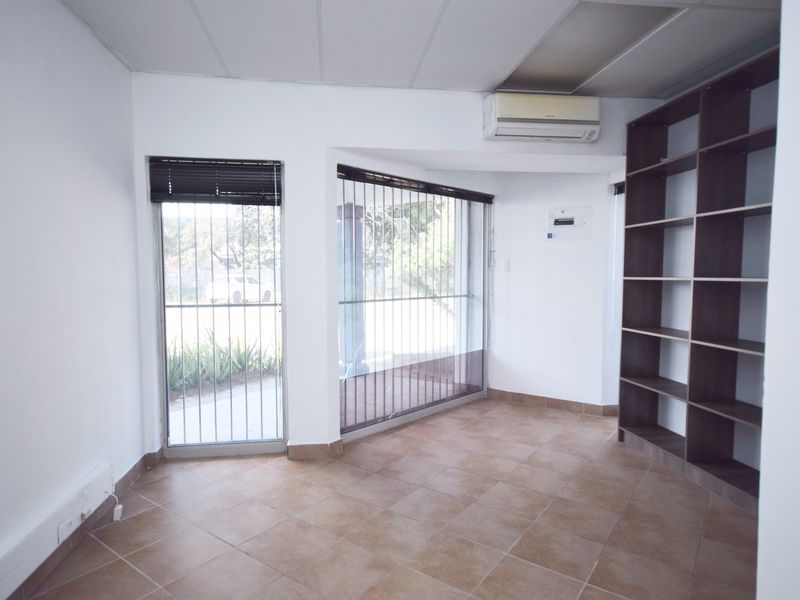 A bright, empty office space featuring tiled flooring, built-in shelving, a wall-mounted air conditioning unit, and large windows with security bars.