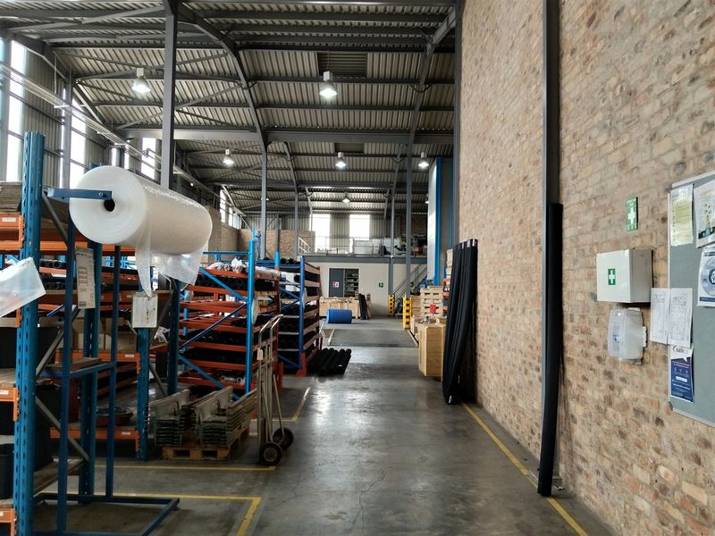 Spacious industrial warehouse featuring exposed brick walls, high steel truss ceilings, concrete flooring, and a mezzanine level, suitable for storage or manufacturing operations.
