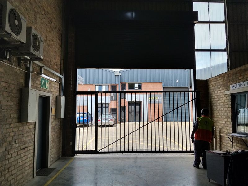 Industrial warehouse space featuring exposed brick walls, concrete flooring, and a large roller shutter door opening to an exterior parking area.