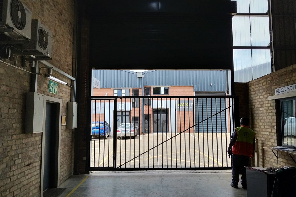 Industrial warehouse space featuring exposed brick walls, concrete flooring, and a large roller shutter door opening to an exterior parking area.