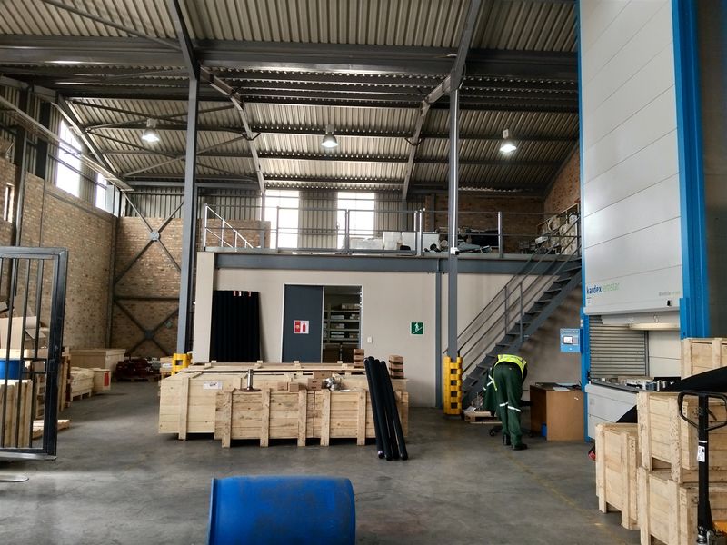 Spacious industrial warehouse featuring high ceilings with exposed steel trusses, a mezzanine level with staircase access, and an integrated automated storage system.