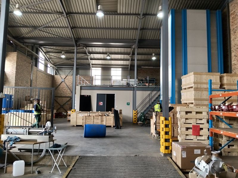Spacious industrial warehouse featuring high ceilings with exposed steel trusses, a mezzanine level, concrete flooring, and brick wall finishes.