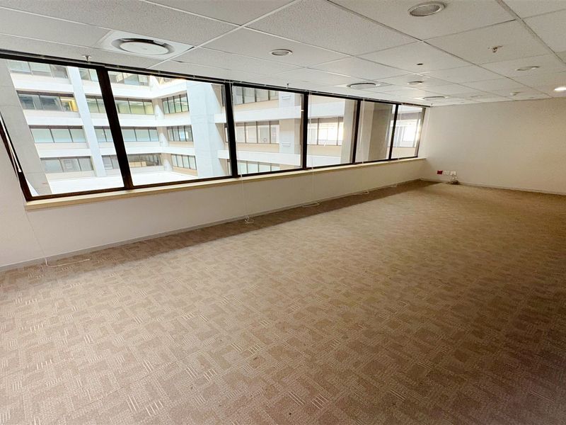 A spacious, empty office suite featuring carpeted flooring, a suspended ceiling with recessed lighting, and large windows overlooking an internal courtyard.