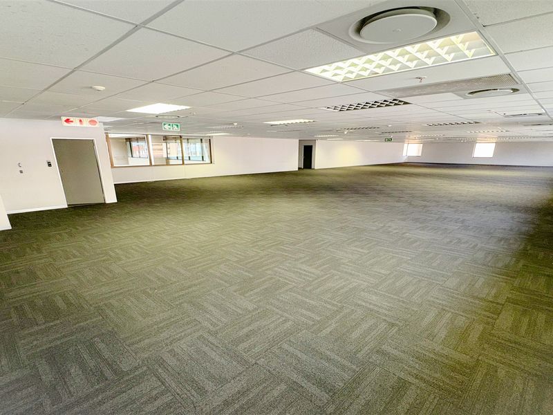 A spacious, open-plan office area featuring carpeted flooring, a suspended ceiling with recessed lighting, and integrated air conditioning vents.