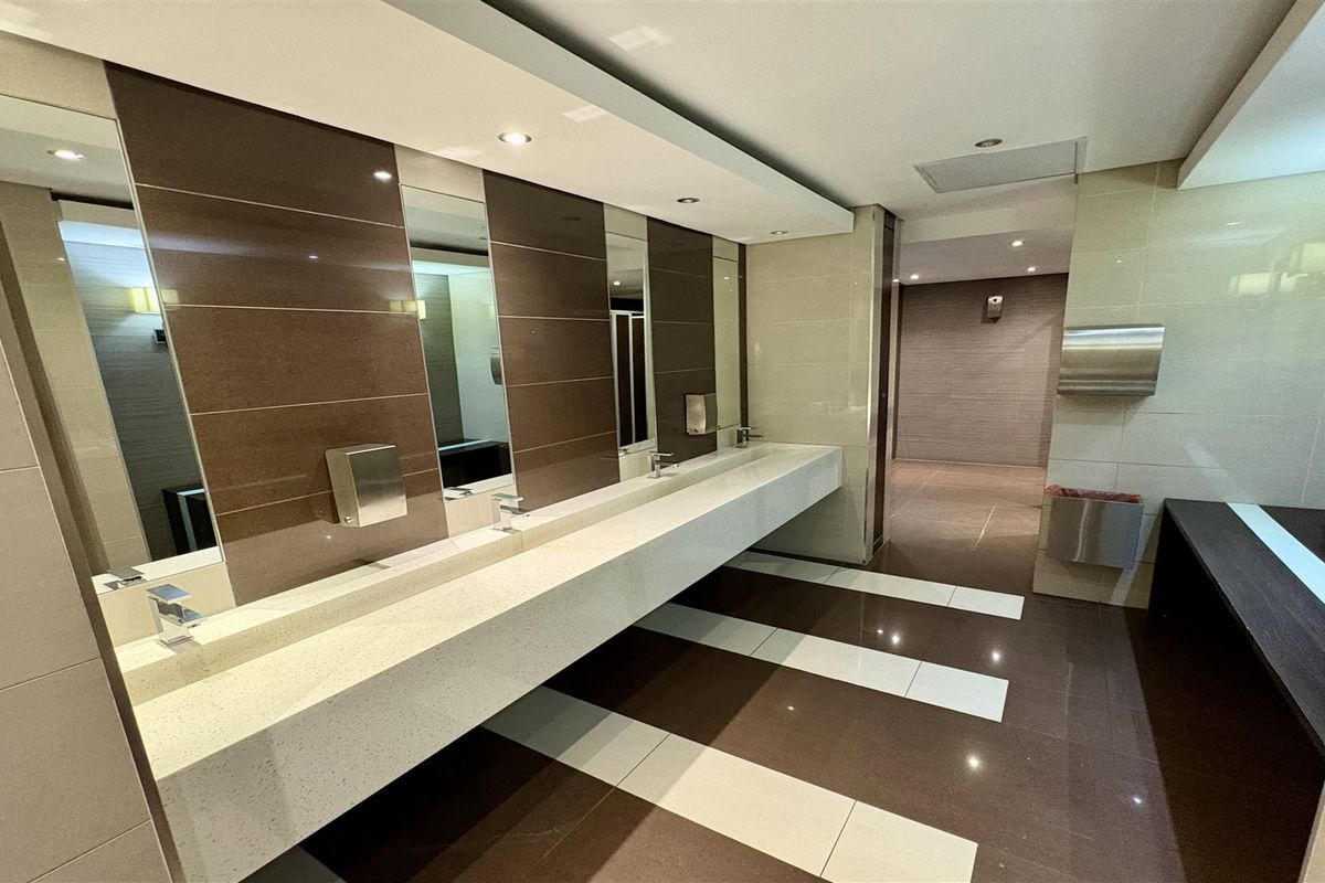 Modern commercial bathroom featuring a long integrated vanity countertop, tiled finishes, and recessed lighting in a clean, well-maintained office facility.