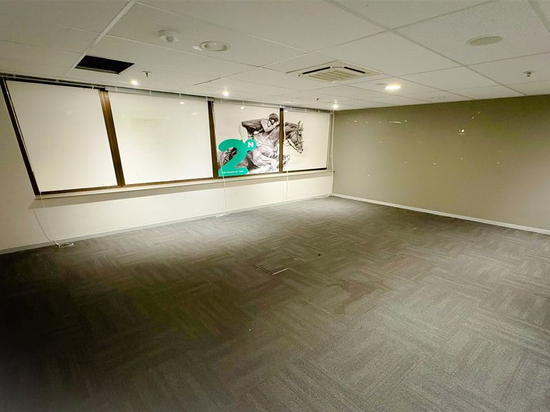 An empty office space featuring carpet tiles, a suspended ceiling with recessed lighting, ducted air conditioning, and large windows with roller blinds.