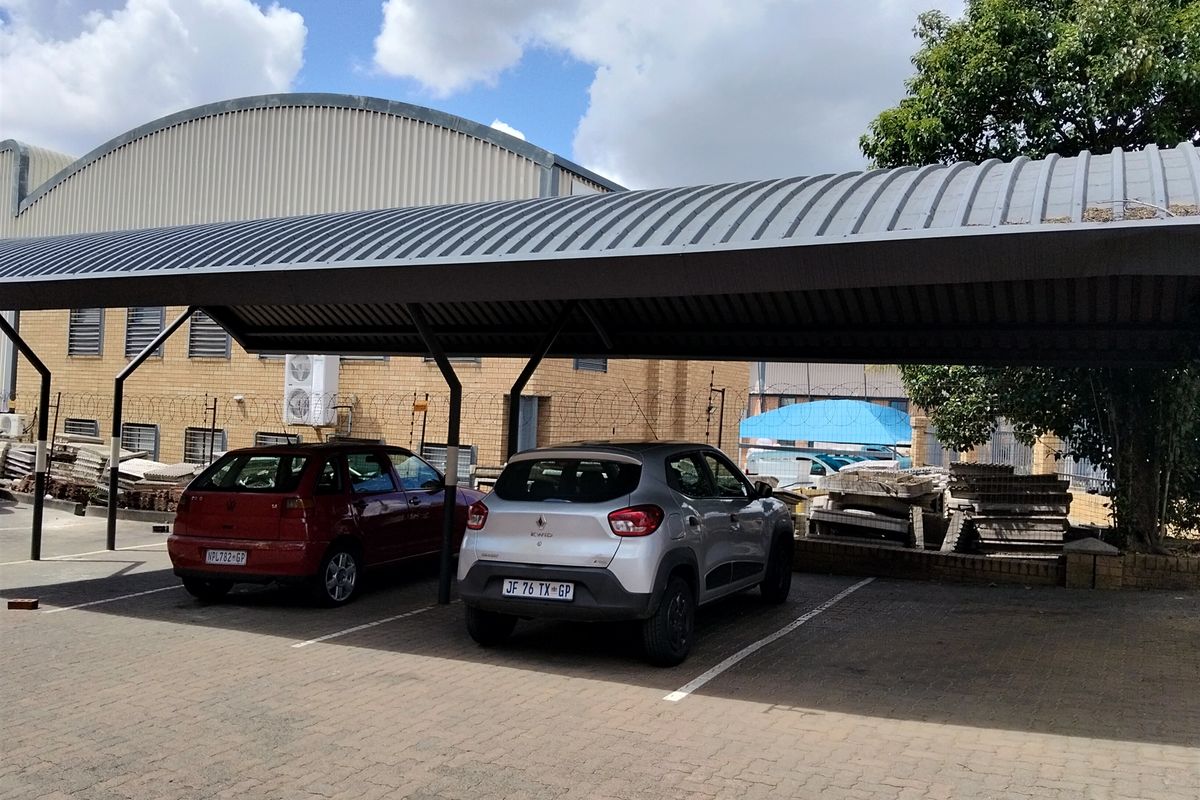 Industrial property exterior featuring a covered parking area with metal roofing and paved bays, adjacent to a brick warehouse structure with security fencing.