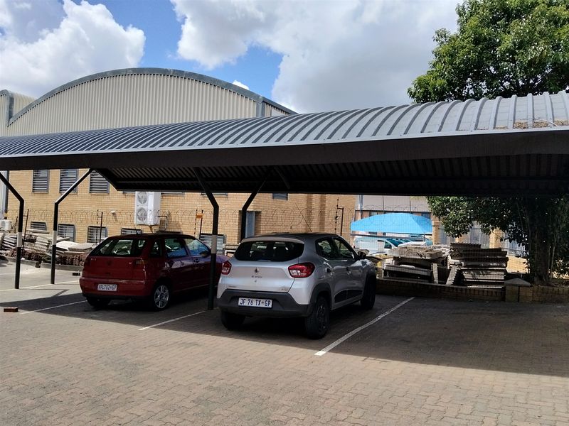 Industrial property exterior featuring a covered parking area with metal roofing and paved bays, adjacent to a brick warehouse structure with security fencing.
