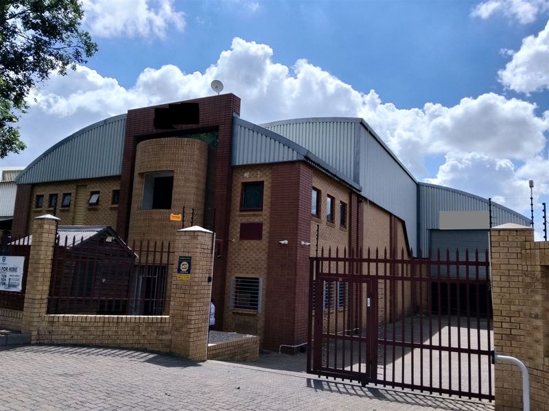 Front elevation of an industrial warehouse facility featuring brick construction, metal roofing, secure perimeter fencing, and a roller shutter access door.