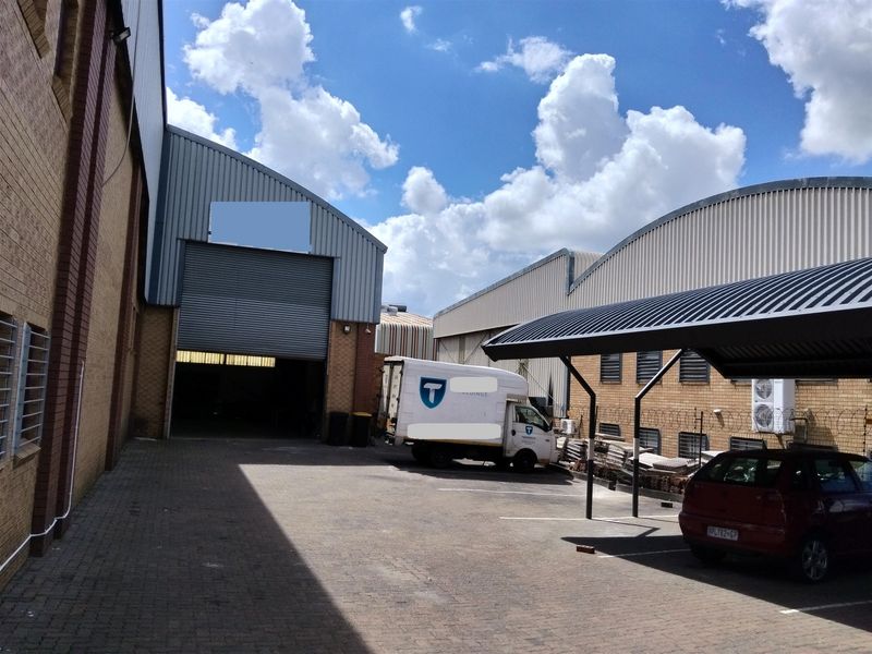 Industrial rear yard featuring a large roller shutter door, paved parking area, and a covered carport structure.