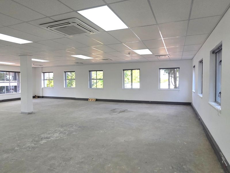 A spacious, open-plan office area featuring a suspended ceiling, recessed lighting, integrated cassette air conditioning, and large windows providing ample natural light.
