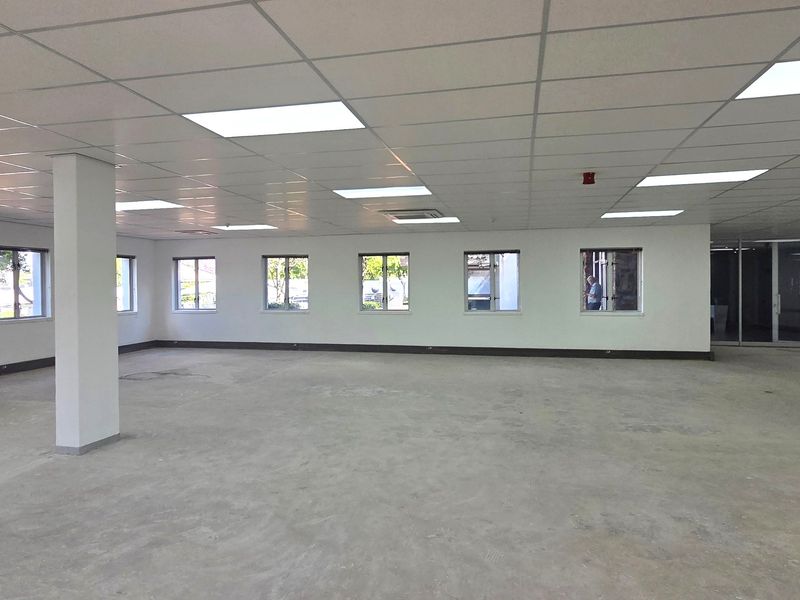 A spacious, open-plan office area featuring a suspended ceiling with recessed lighting, central air conditioning, and large windows, currently awaiting floor finishing.