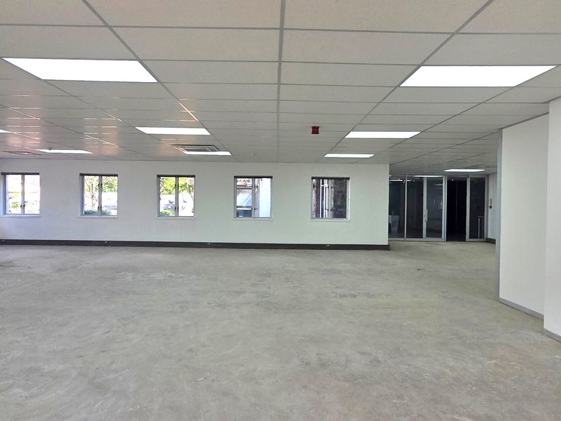 A spacious, open-plan office area featuring a suspended ceiling with recessed lighting, large windows, and glass partition doors, currently awaiting final flooring installation.
