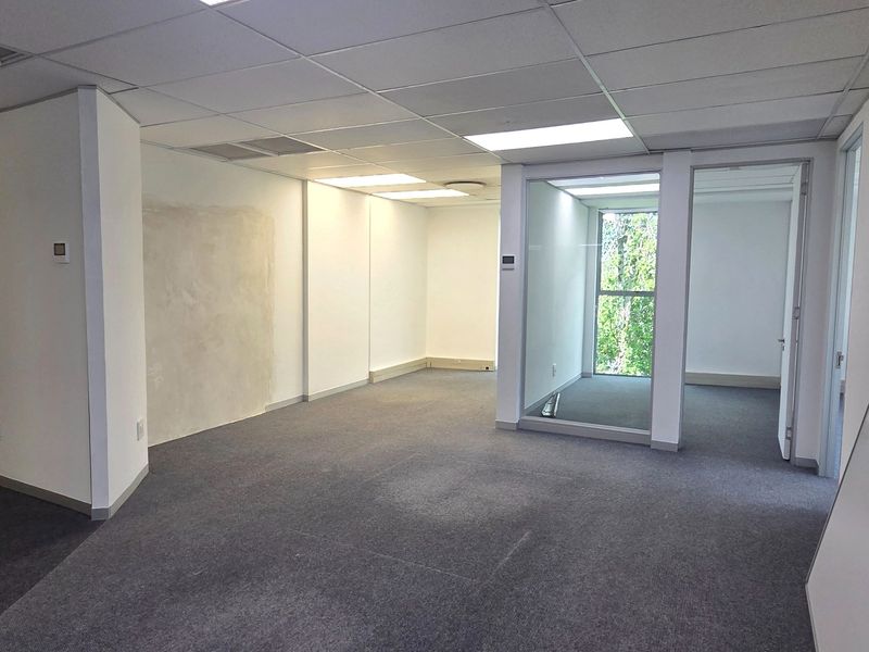 An empty, open-plan office space featuring carpeted flooring, a suspended ceiling with recessed lighting, and a glass partition wall.