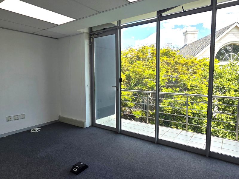 A bright, empty office space featuring carpeted flooring, recessed lighting, and a glass sliding door leading to a private balcony with leafy views.