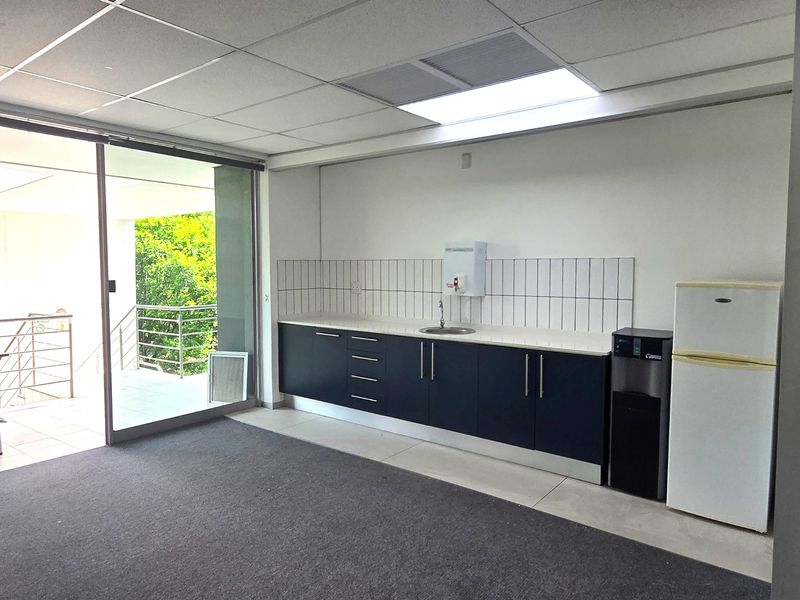 A functional office kitchenette featuring dark cabinetry, a sink, tiled backsplash, and direct access to an outdoor balcony area.
