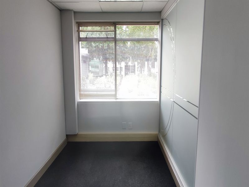 A compact, empty office space featuring a suspended ceiling, carpeted flooring, and a frosted glass partition wall with natural light from a window.