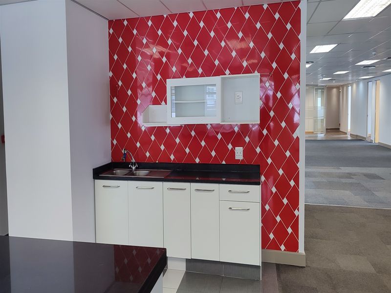 A compact office kitchenette featuring white cabinetry, a dark countertop, stainless steel sink, and a distinctive red and white diamond-patterned tiled backsplash.