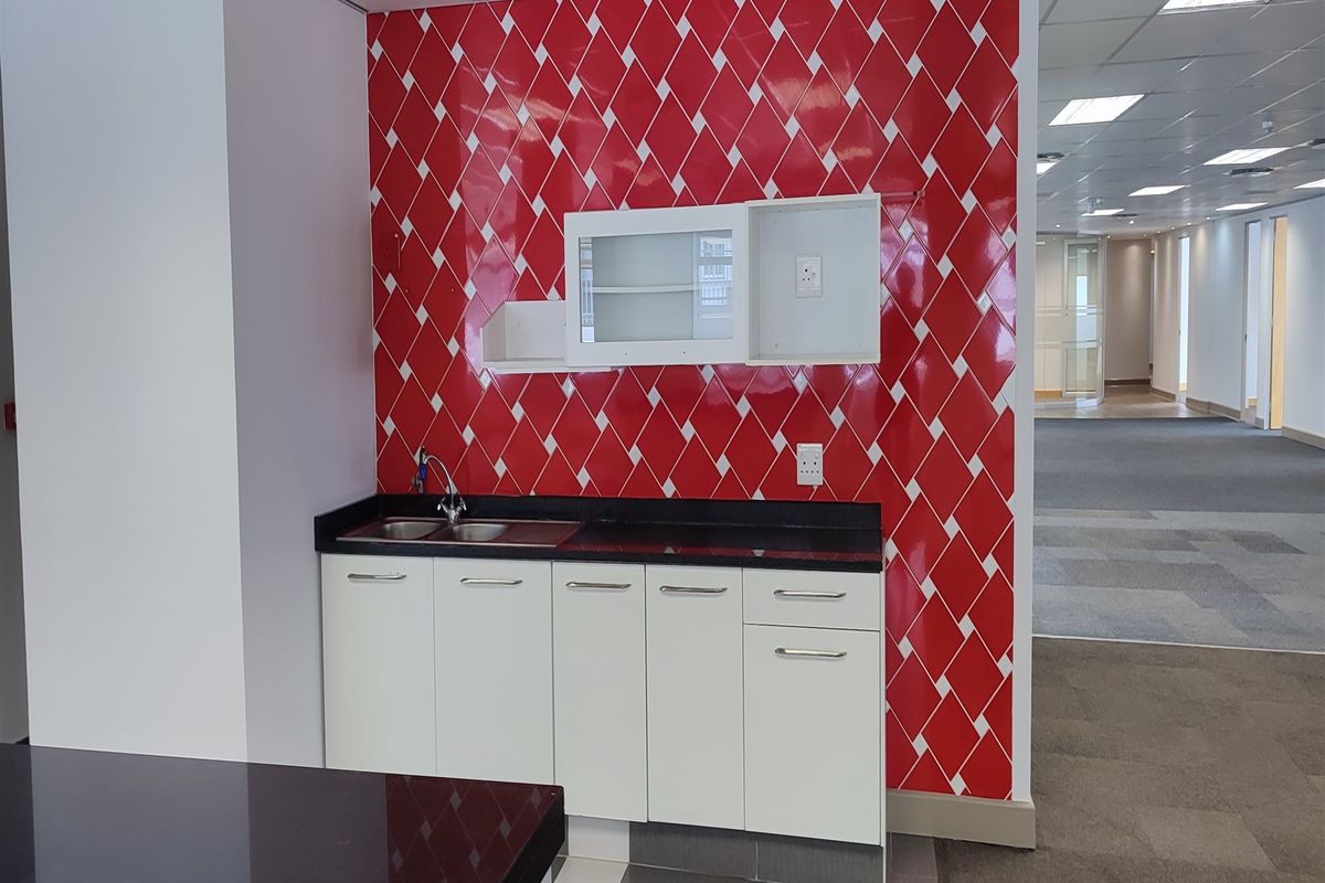 A compact office kitchenette featuring white cabinetry, a dark countertop, stainless steel sink, and a distinctive red and white diamond-patterned tiled backsplash.