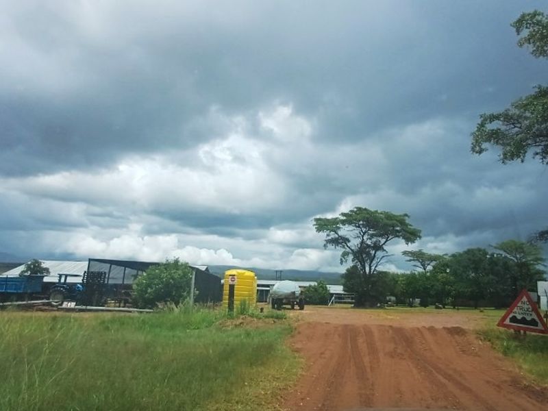A wide-angle view of a farm entrance featuring a dirt driveway, water storage infrastructure, and surrounding agricultural outbuildings under a cloudy sky.
