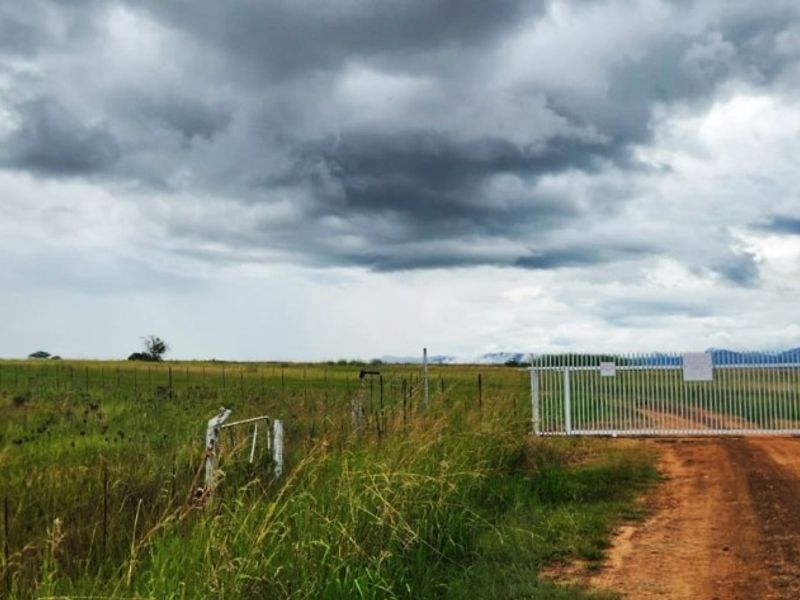 A view of the farm entrance featuring a white metal security gate, wire fencing, and a dirt access road leading into the open landscape.