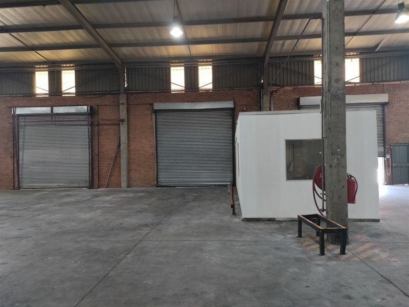 Spacious industrial warehouse featuring high ceilings, multiple roller shutter doors, exposed brickwork, concrete flooring, and a dedicated internal office enclosure.