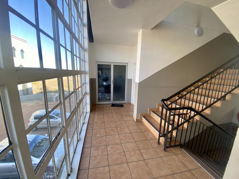 A bright, tiled hallway featuring a staircase with metal railings and large steel-framed windows providing natural light and exterior views.