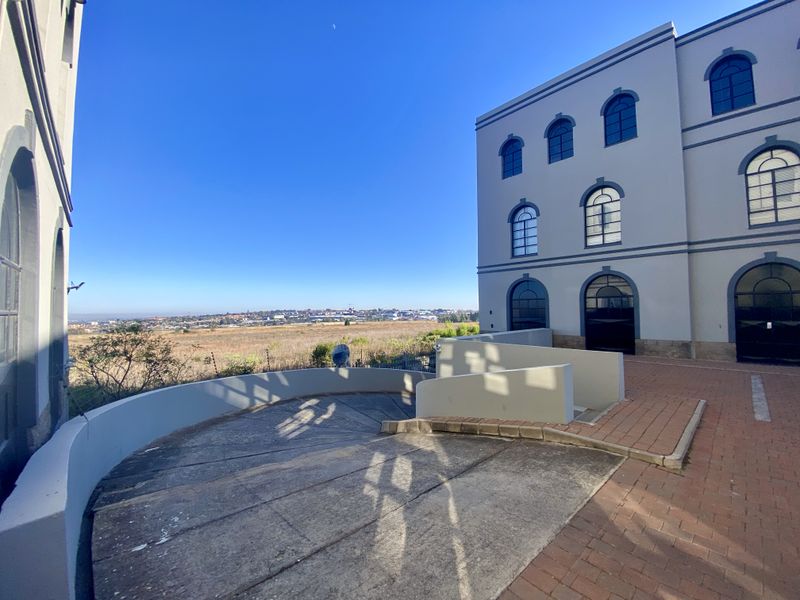 Exterior view of a multi-story industrial building featuring arched windows, a paved driveway, and a retaining wall with surrounding landscape views.