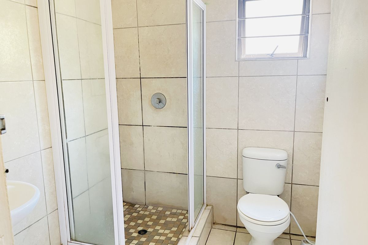A functional bathroom featuring a glass-enclosed shower, a standard toilet, and neutral wall and floor tiling with a small window for ventilation.