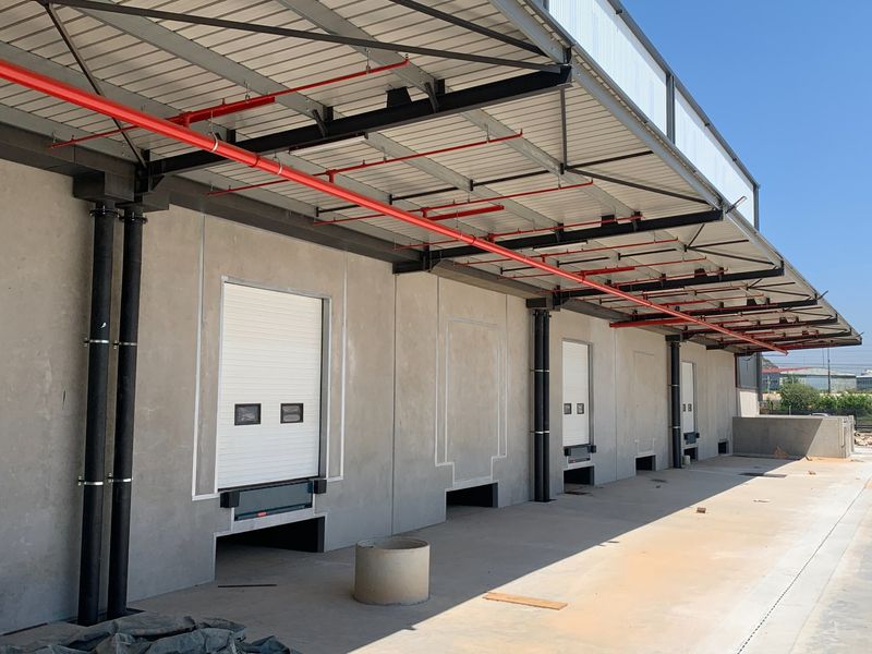 Industrial loading bay area featuring multiple roller shutter doors, overhead fire sprinkler system, and a durable concrete apron under a steel roof structure.