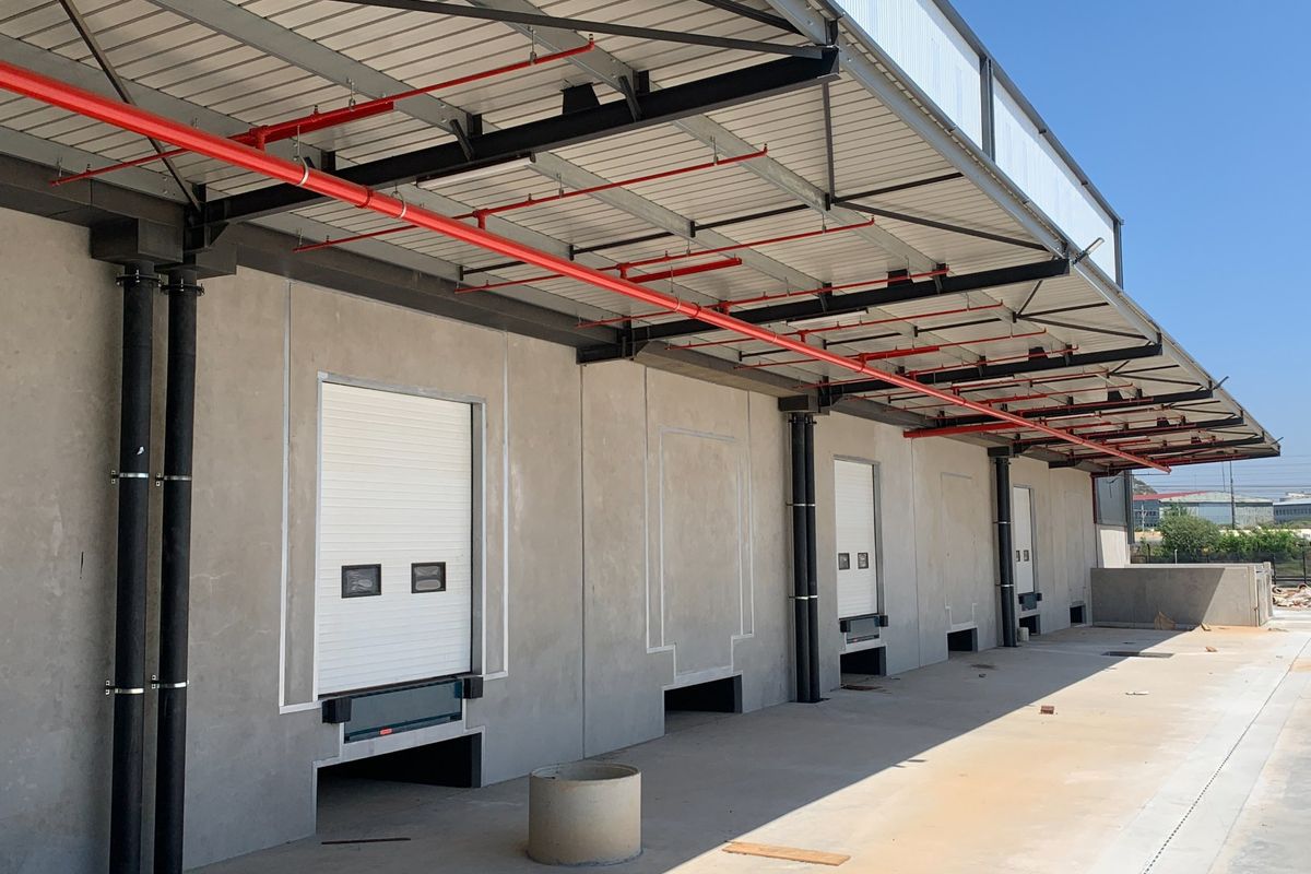 Industrial loading bay area featuring multiple roller shutter doors, overhead fire sprinkler system, and a concrete loading apron.