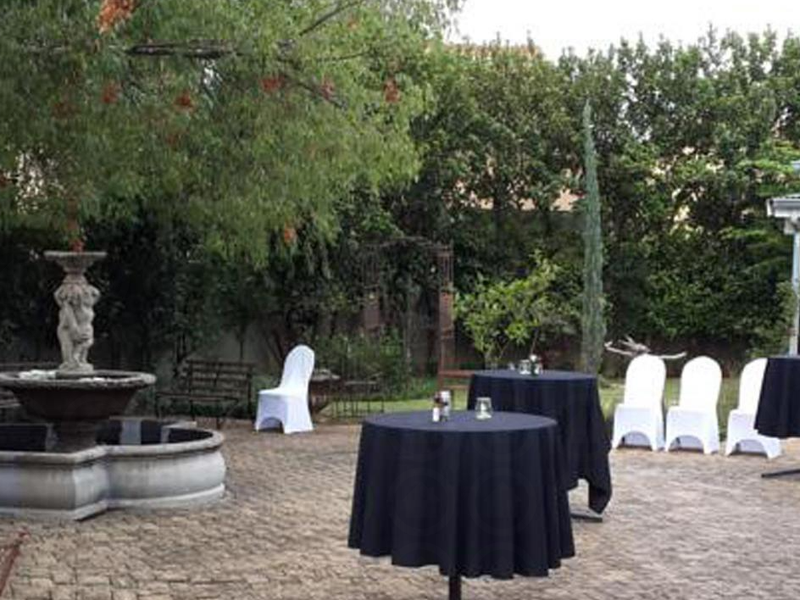 A spacious paved outdoor patio area featuring a decorative stone fountain, mature trees, and a covered veranda entrance to the guest house.