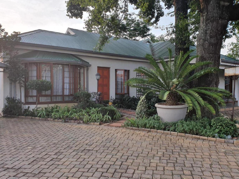 Front elevation of a guest house featuring a paved driveway, established garden, bay window, and a metal roof.