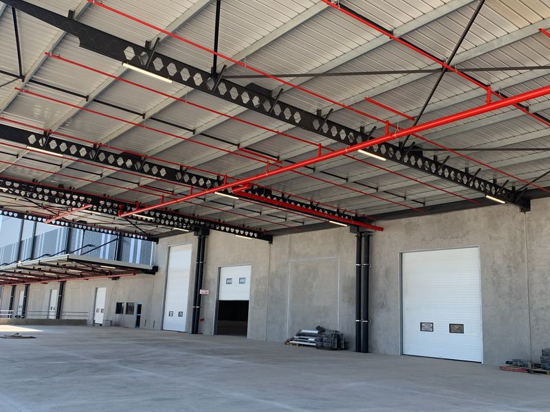 Spacious industrial warehouse interior featuring high ceilings with exposed steel trusses, fire sprinkler systems, and multiple large roller shutter doors for efficient loading access.