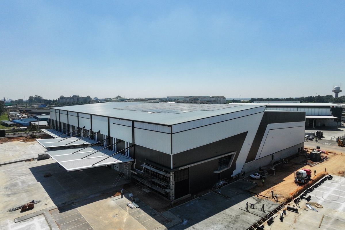 Aerial view of a modern industrial warehouse facility featuring a large metal roof with solar panels, loading bay canopies, and extensive concrete hardstand areas.