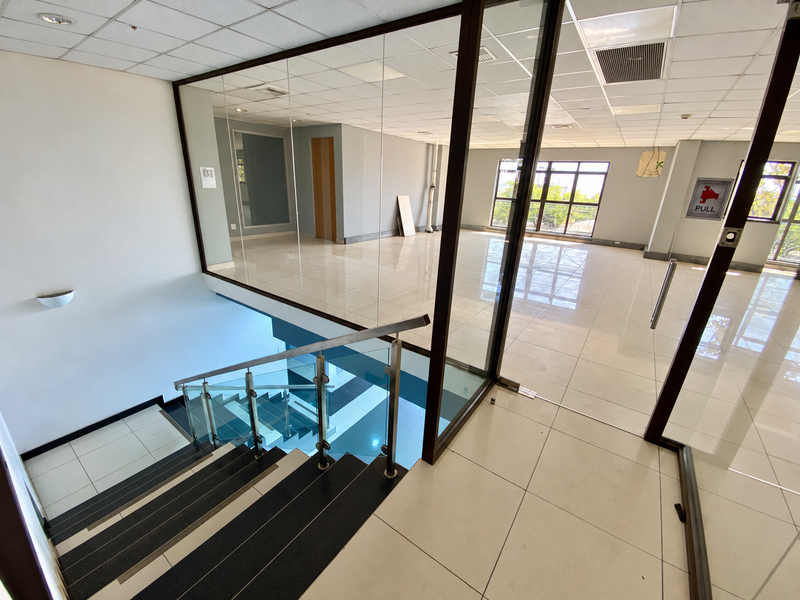 Spacious industrial office hallway featuring glass partitions, tiled flooring, and a modern staircase with a glass balustrade, offering a bright and professional workspace.