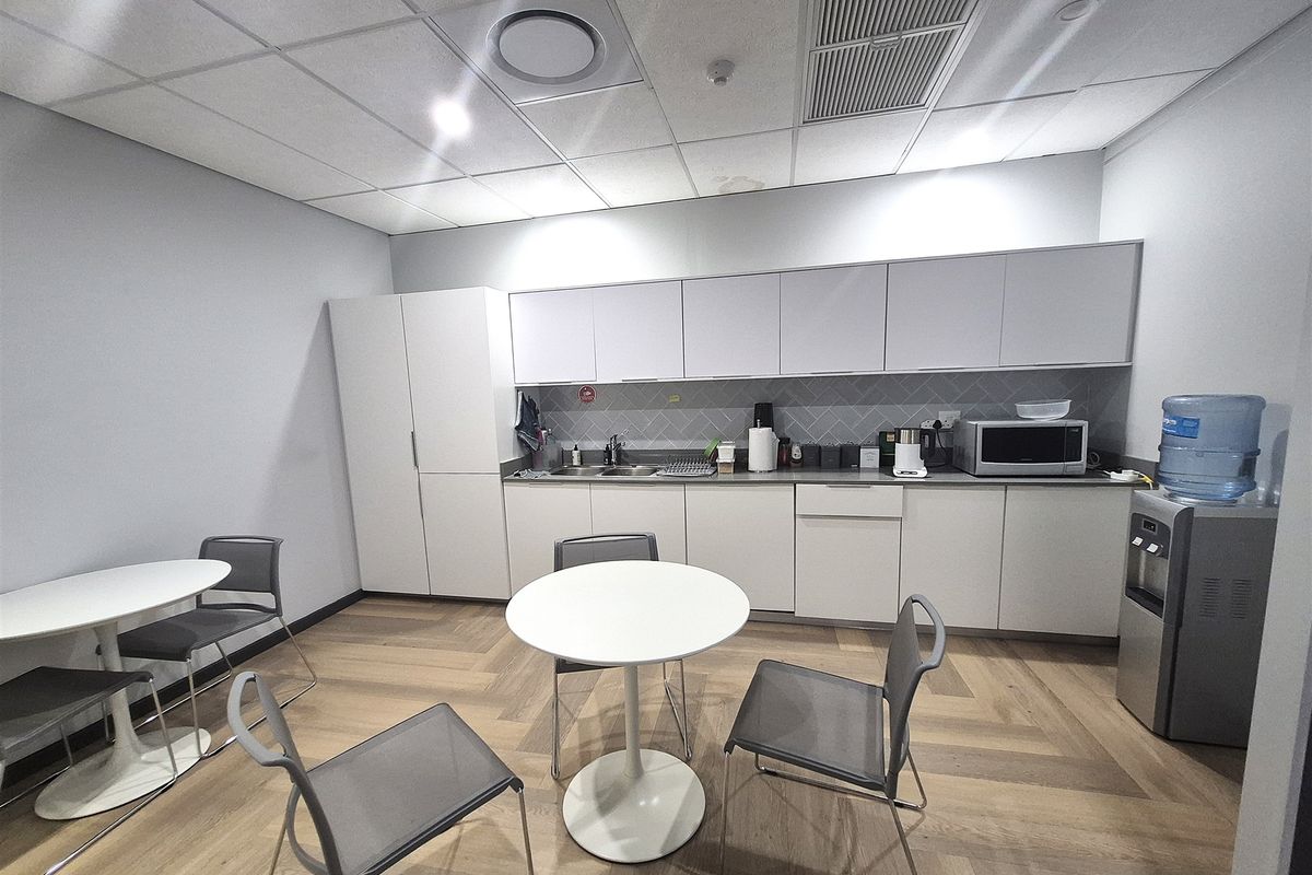 A modern office kitchenette featuring clean white cabinetry, a stainless steel sink, herringbone flooring, and a suspended ceiling with integrated climate control.
