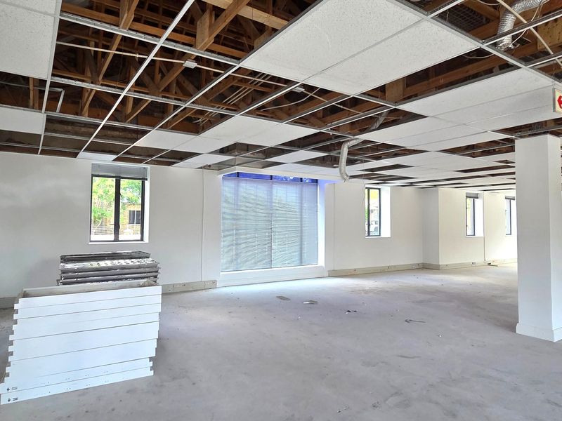 An open-plan office space currently undergoing renovations, featuring an exposed ceiling structure, large windows with blinds, and a blank canvas interior.