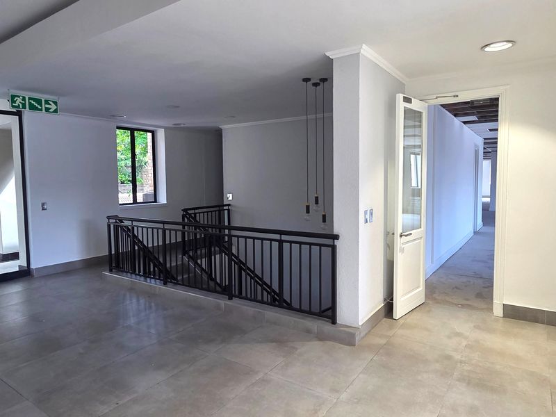 A bright and spacious office hallway featuring modern tiled flooring, a metal staircase railing, and recessed ceiling lighting with access to adjacent office rooms.