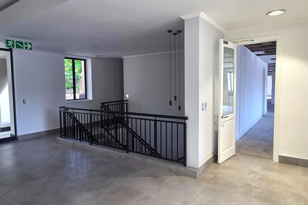 A bright and spacious office hallway featuring modern tiled flooring, a metal staircase railing, and recessed ceiling lighting with access to adjacent office rooms.