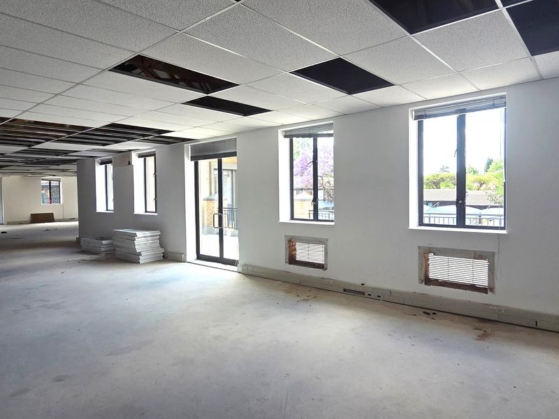 An open-plan office space in shell condition, featuring a suspended ceiling with missing tiles, large windows, and an unfinished floor.
