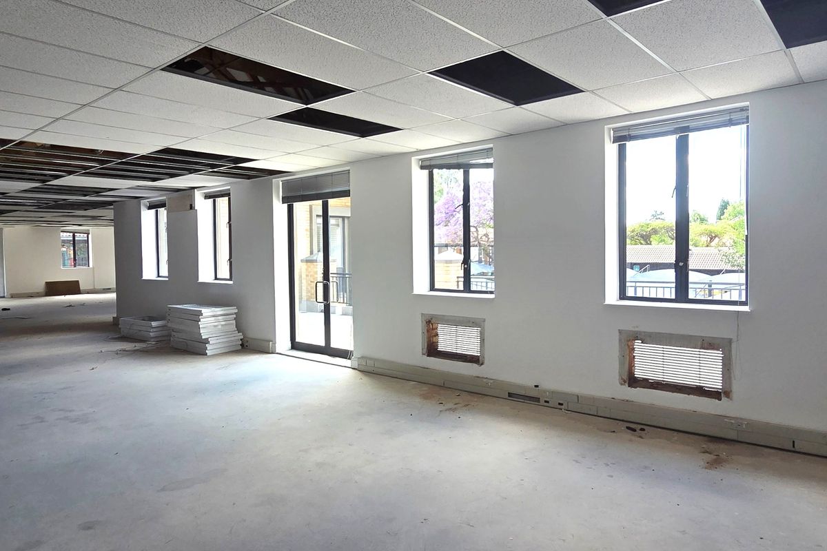 An open-plan office space in shell condition, featuring a suspended ceiling with missing tiles, large windows, and an unfinished floor.