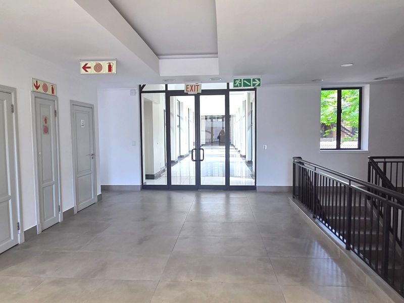 A spacious, well-lit office hallway featuring tiled flooring, a metal balustrade, and glass-panelled double doors leading to an adjacent corridor.