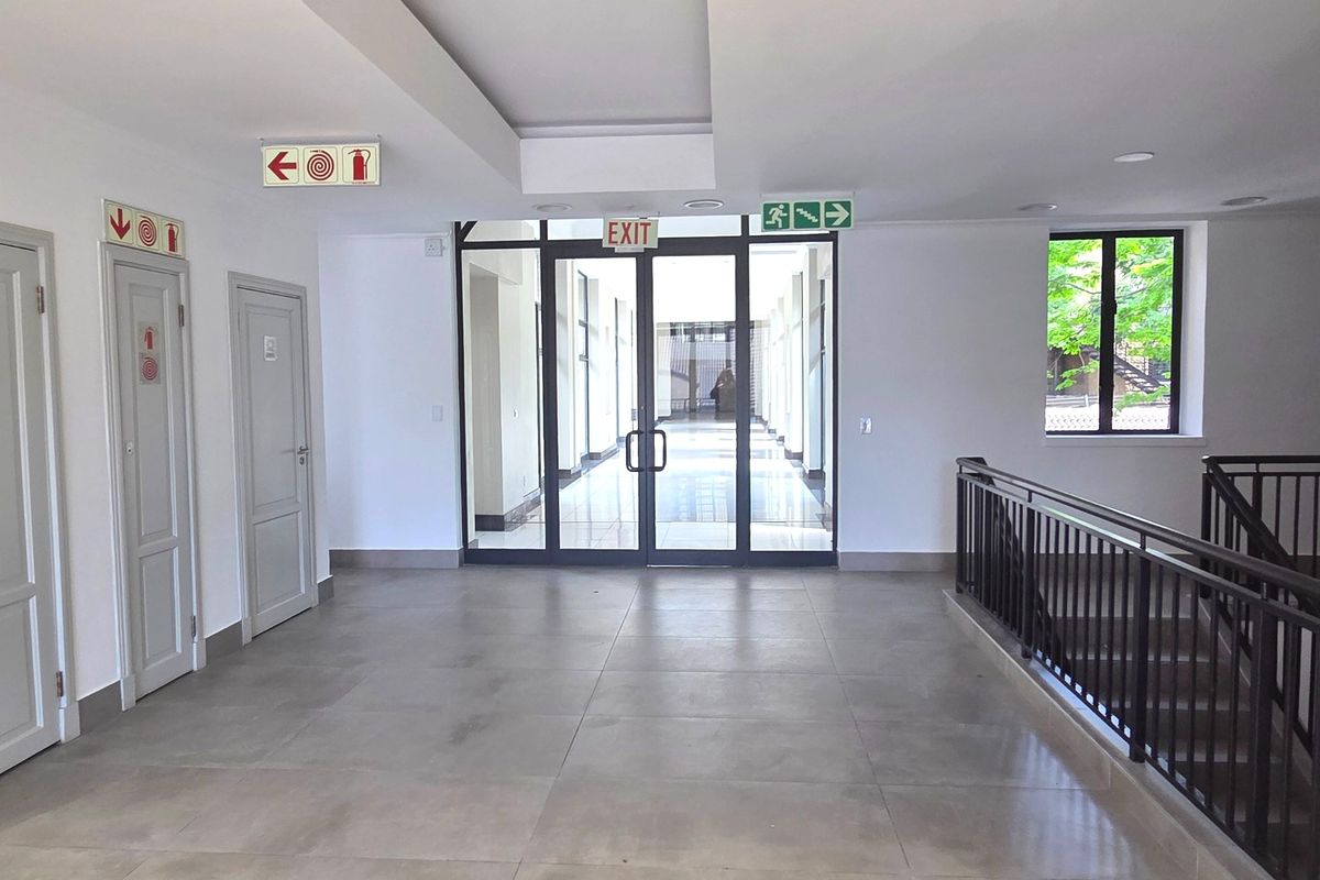 A spacious, well-lit office hallway featuring tiled flooring, a metal balustrade, and glass-panelled double doors leading to an adjacent corridor.