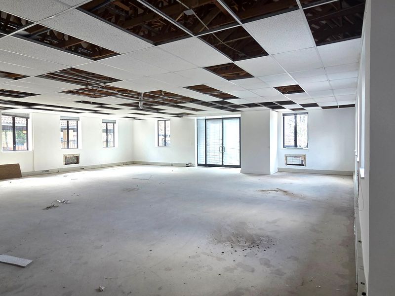Spacious open-plan office area featuring large windows, sliding glass doors, and a suspended ceiling grid currently undergoing renovation with missing tiles.