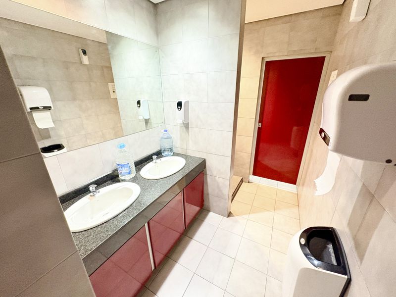 A functional office bathroom featuring a double vanity with integrated sinks, large wall-mounted mirror, and neutral tiled finishes throughout.