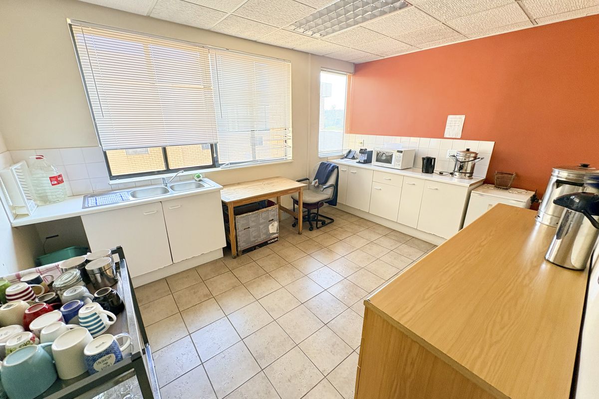 Functional office kitchenette featuring double basin sink, laminate cabinetry, tiled flooring, and ample natural light through large windows with venetian blinds.